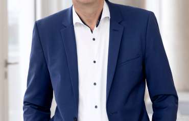 Dr. Marc Kaninke Ein Mann mit kurzem grauem Haar und Brille steht in einem modernen, hellen Innenraum. Er trägt ein dunkelblaues Sakko mit passender Hose, ein weißes Hemd mit dunklen Knöpfen und hat die Hände in den Hosentaschen. Im Hintergrund sind große Fenster und helle Wände zu sehen.