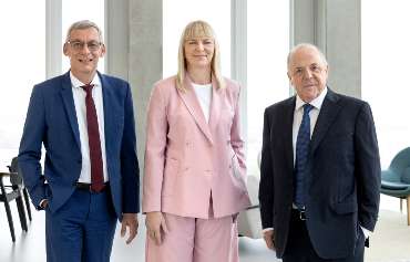 Der Vorstand der Landwirtschaftlichen Rentenbank Drei Personen stehen nebeneinander in einem modernen, hellen Raum mit großen Fenstern. Die Person links trägt einen blauen Anzug mit weißem Hemd und roter Krawatte, die Person in der Mitte einen rosafarbenen Hosenanzug mit weißem Oberteil, und die Person rechts einen dunkelblauen Anzug mit weißem Hemd und dunkelblauer Krawatte. Im Hintergrund sind einige Stühle und ein Tisch zu sehen.