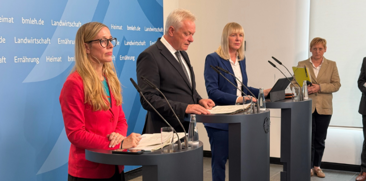 m Vordergrund stehen drei Personen hinter Pulten, die mit Mikrofonen ausgestattet sind. Die mittlere Person trägt einen dunklen Anzug und hält ein Manuskript, während die beiden anderen Personen links und rechts jeweils einen farbigen Blazer tragen. Im Hintergrund befindet sich eine weitere Person mit Tablet. Die Wand hinter den Pulten zeigt das Logo und die Schriftzüge des Bundesministeriums für Ernährung und Landwirtschaft. Die Szene vermittelt den Eindruck einer Pressekonferenz zum Erntebericht, bei der Alois Rainer und Nikola Steinbock im Mittelpunkt stehen.
